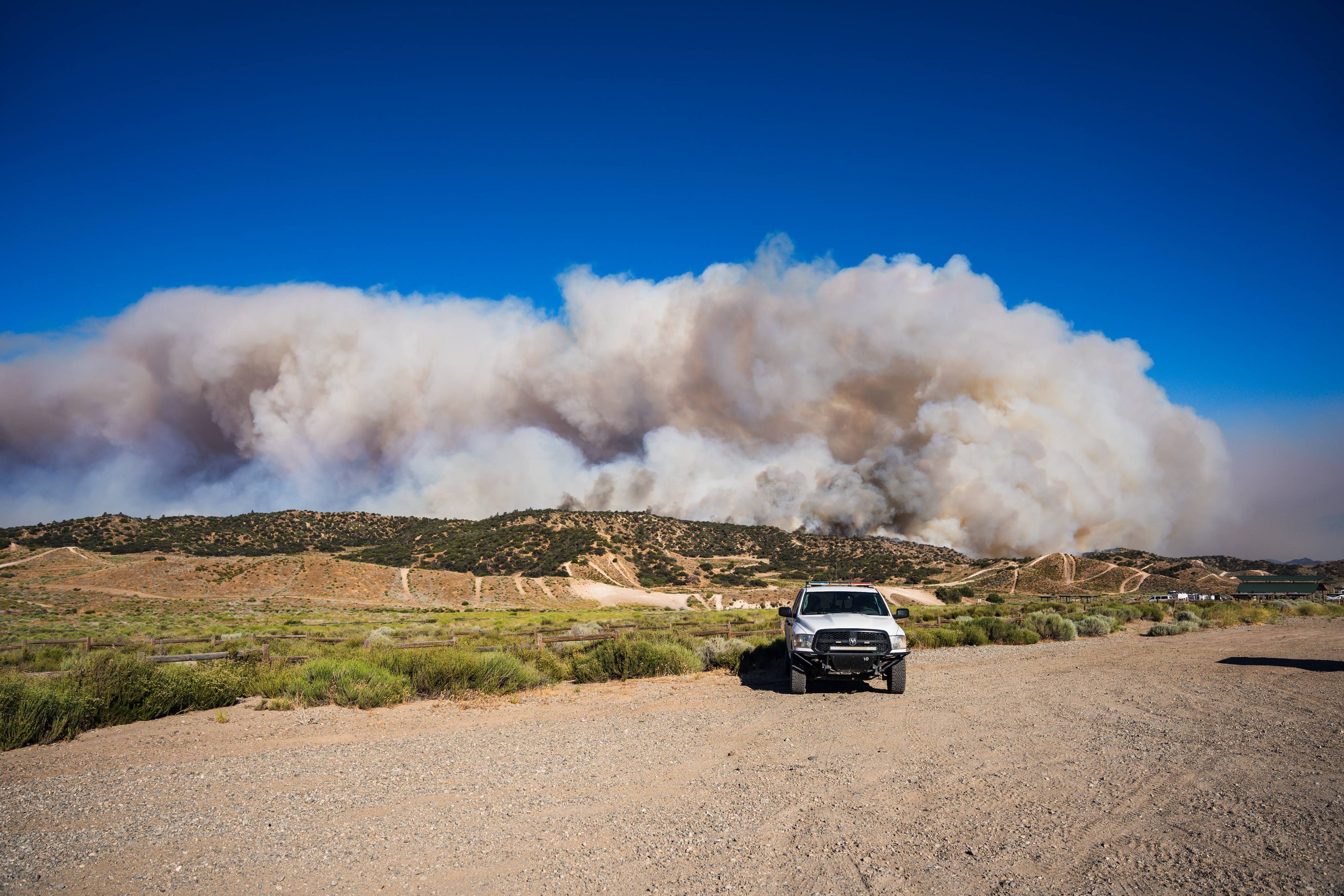 California State Parks Ranger in a white truck parked on a dirt road assists visitors in evacuating the park as plumes of smoke from the Post Fire can be seen within Hungry Valley SVRA.