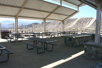 Picnic eating area under the metal shade structure with many picnic tables and food preparation area