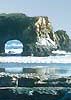 Ocean shot with enormous rock jutting from water, which has a large hole/tunnel carved out from the bottom