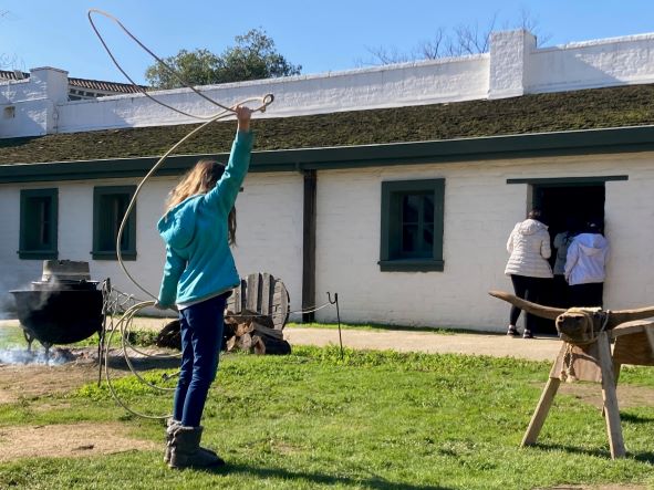 A child wearing jeans and a sweatshirt turns a rope lariat over her head aiming her throw to rope a wooden bulls head mounted to a sawhorse