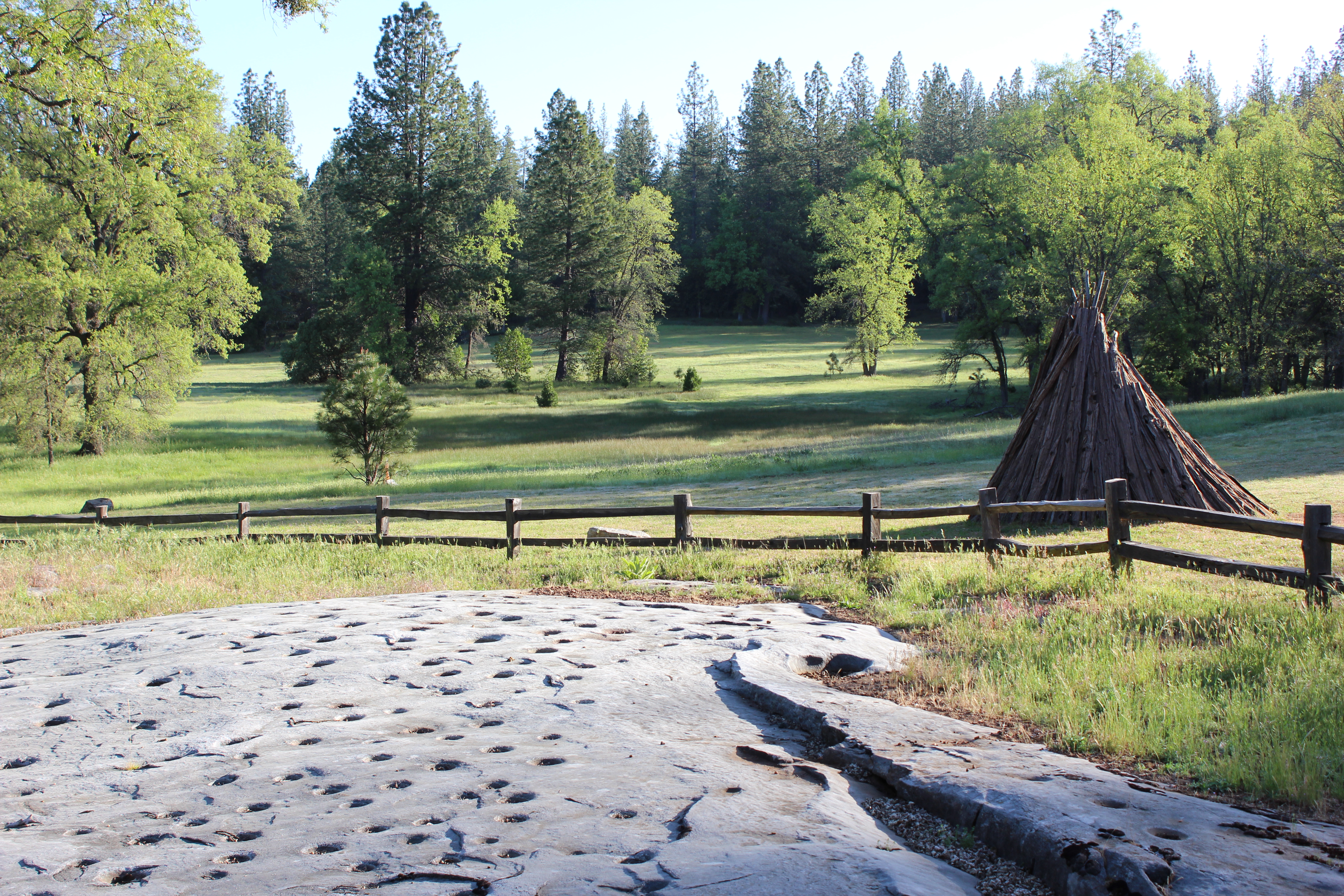Grinding rock with meadow and bark house in the background. 