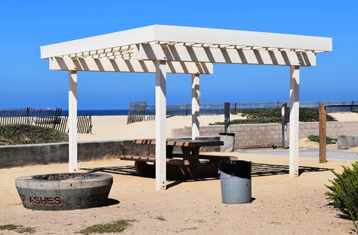 Ramada with a picnic bench and a fire ring sit on the sand.